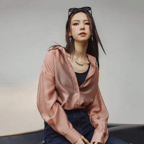 Woman wearing a pink shirt and dark pants with sunglasses on her head, sitting against a neutral background.